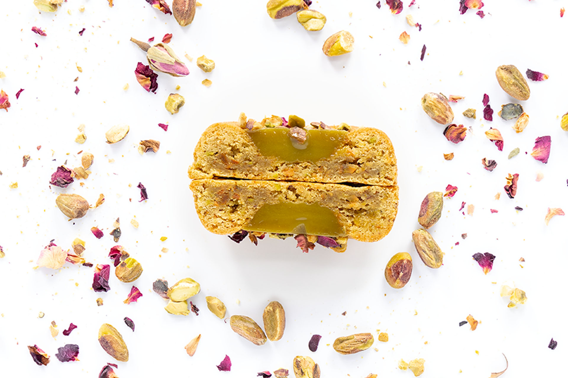 A pistachio macaron with a creamy filling, surrounded by scattered pistachio nuts, espresso beans, and rose petals on a white background.