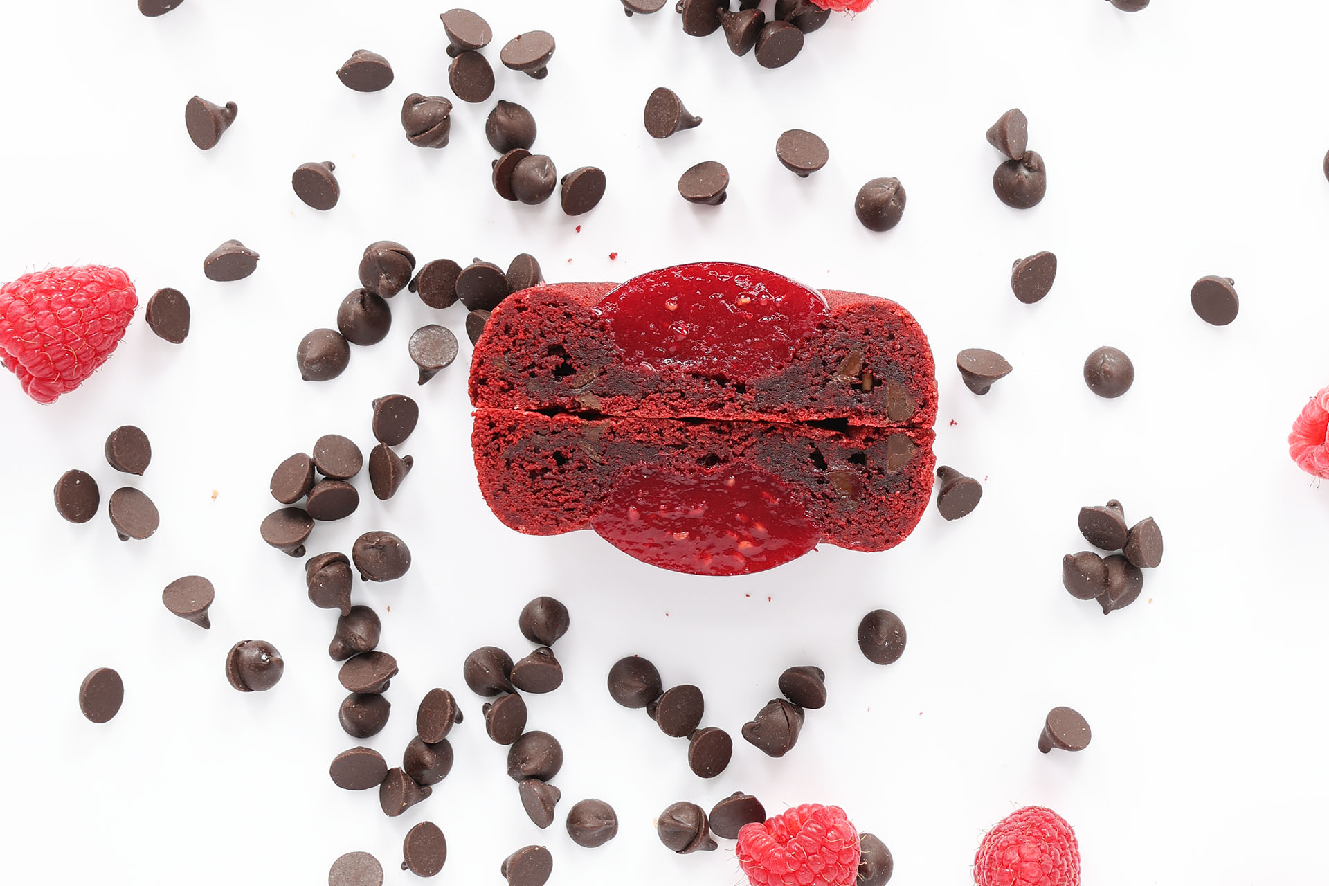 A red macaron with a chocolate and espresso filling, surrounded by scattered chocolate chips and raspberries on a white surface.