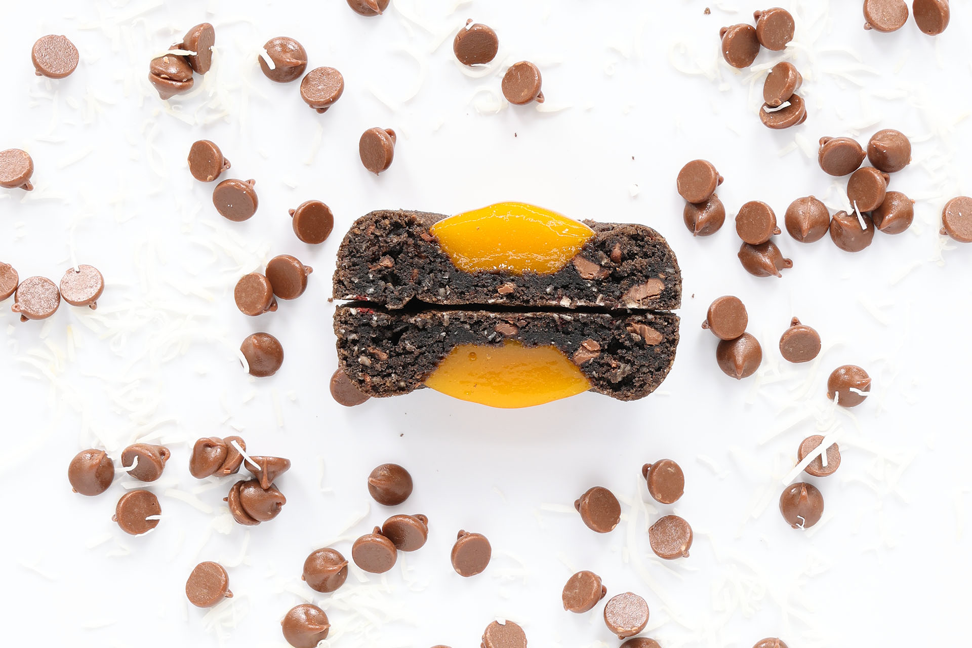 Chocolate cookie filled with apricot jam, surrounded by chocolate chips, espresso flakes, and coconut flakes on a white surface.