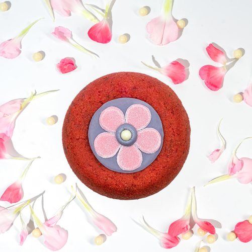 A red velvet cake with a decorative edible flower in the center, surrounded by scattered pink petals and golden beads, featured on the homepage, on a white background.