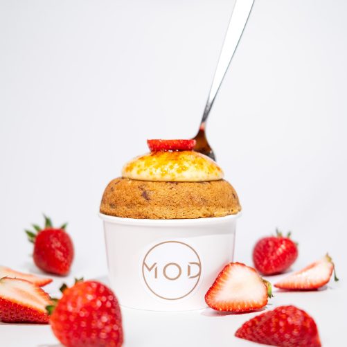 A freshly baked muffin in a white cup with a spoon, topped with cream and a slice of strawberry, surrounded by strawberry halves on a white surface, perfect for homepage SEO.
